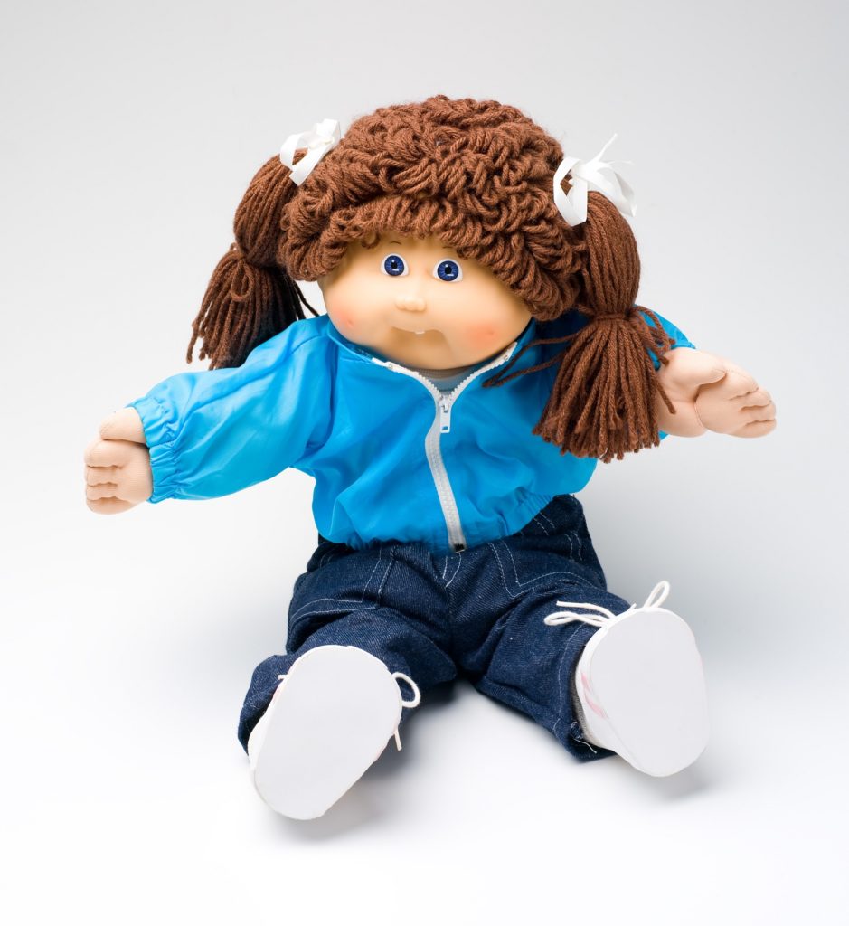 The Cabbage Patch doll has long brown hair tied into pigtails on either side of her head. The doll is dressed a light blue zip-up windcheater and dark blue denim jeans with white shoes. She has her arms out and is in a seated position.